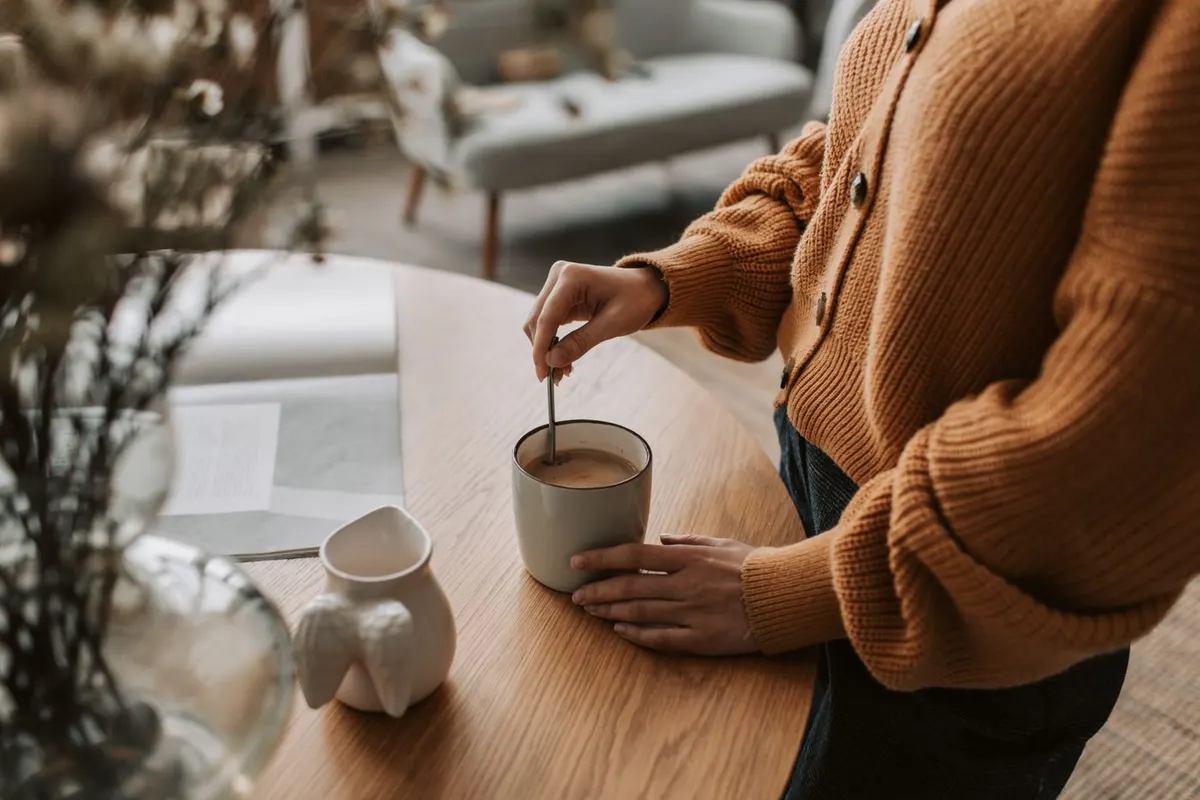 Woman in an orange cardigan stirring a mug of coffee at a wooden table