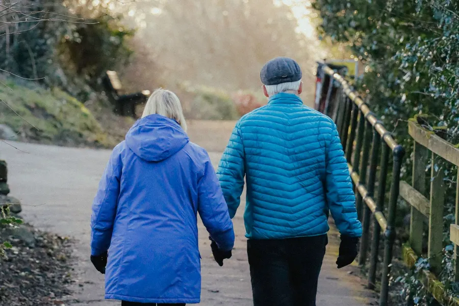 An older couple walking together along a tree-lined path in the winter sun