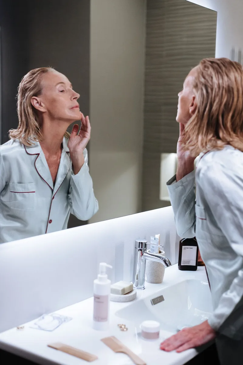 Woman over 50 in pajamas touching her jawline while looking at her skin in a bathroom mirror