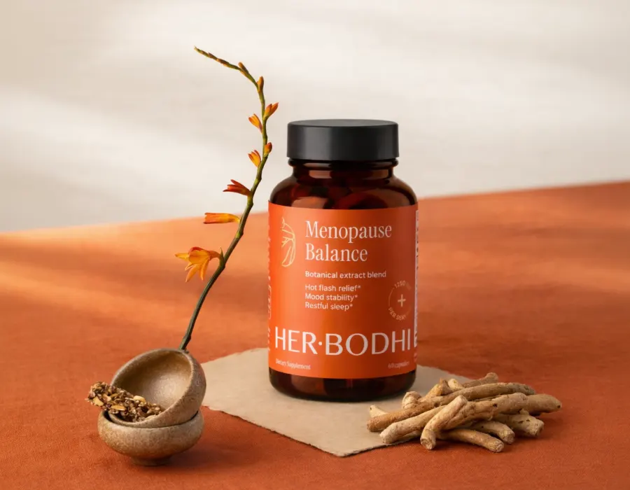 Bottle of Her Bodhi Menopause Balance next to dried ashwagandha root and a small ceramic bowl of herbs