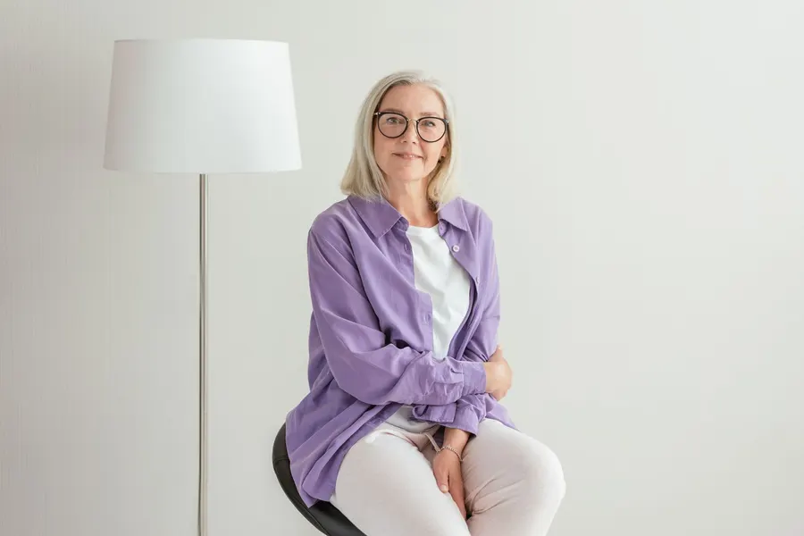 Woman in her fifties with gray hair and glasses sitting calmly in a bright room