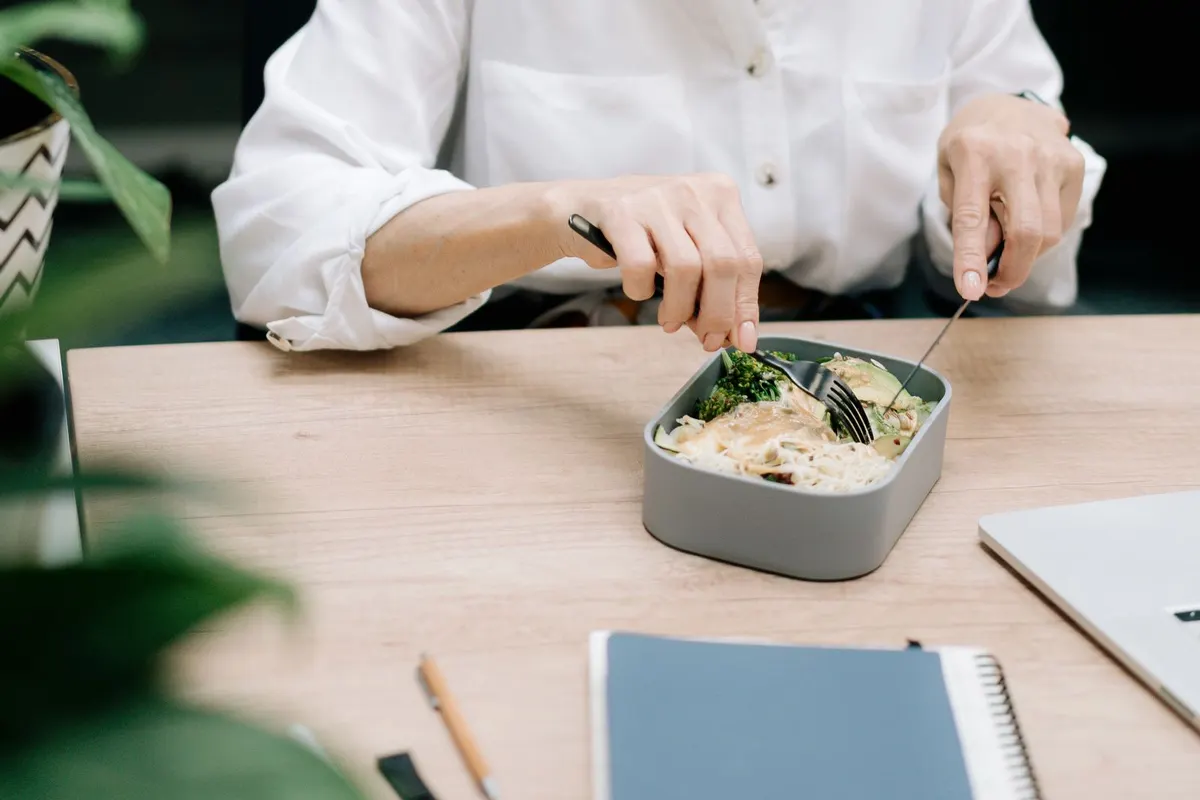 Woman in a white shirt eating a packed lunch of vegetables and grains at a wooden table