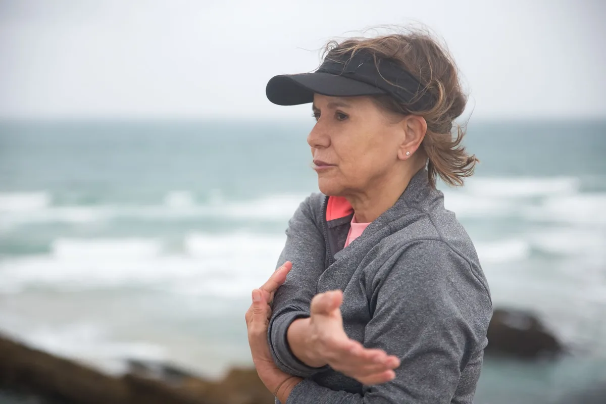 Woman over 50 in a gray jacket and visor stretching one arm across her chest by the sea