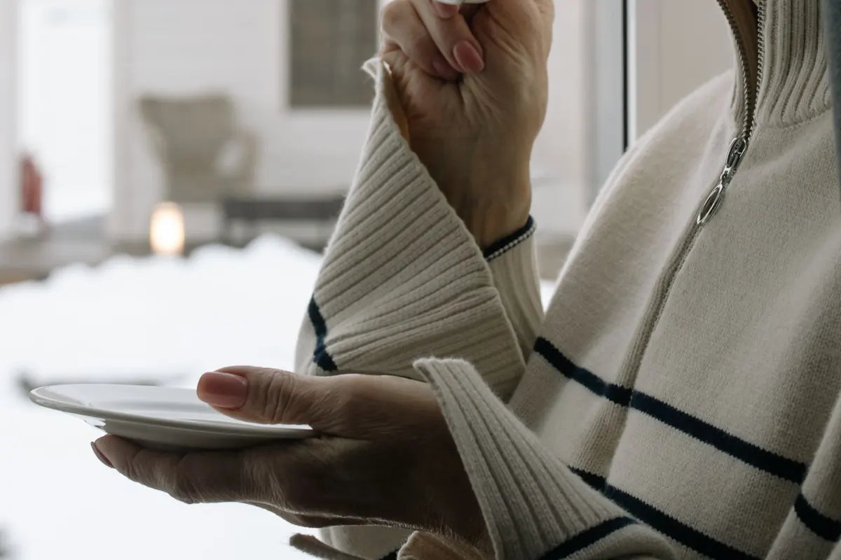 Woman over 50 with gray hair sipping tea from a white cup beside a bright window