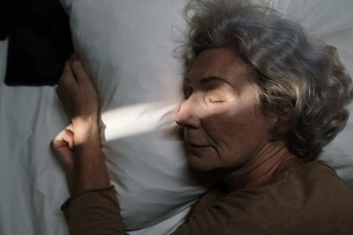 Woman over 50 with gray curly hair sleeping peacefully on a white pillow with soft sunlight across her face