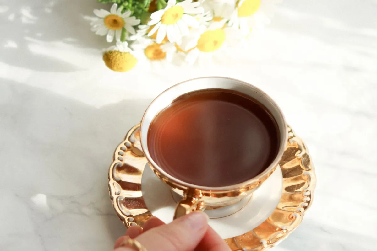 Hand holding a gold-rimmed cup of herbal tea on a saucer next to fresh chamomile flowers