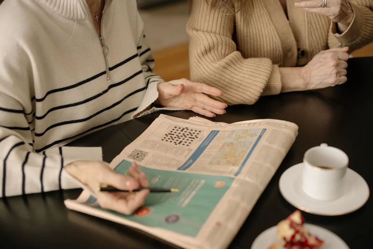 Close-up of a hand holding a pen over a newspaper crossword puzzle next to a cup of coffee