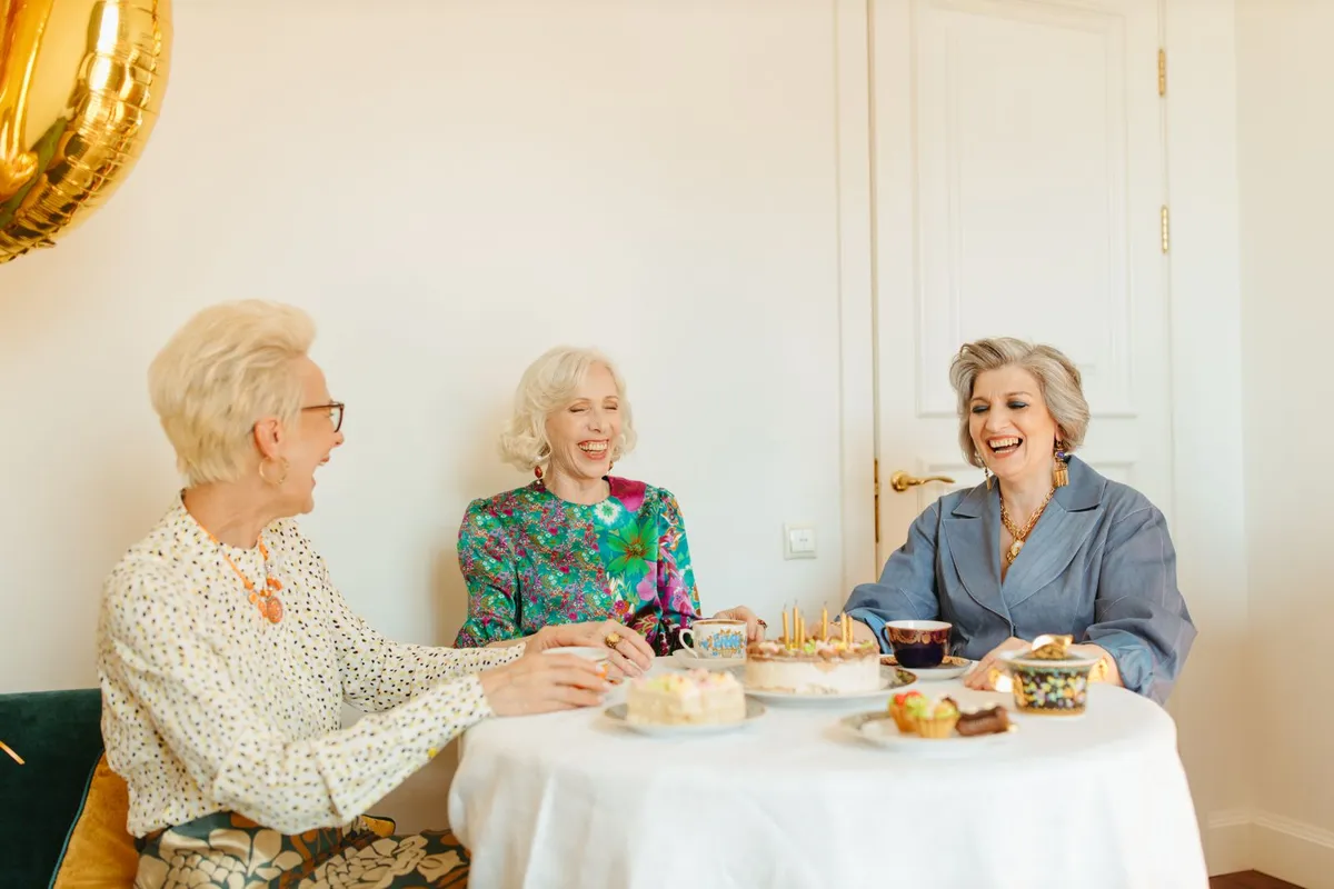 Three women over 50 laughing together over tea and cake at a table at home