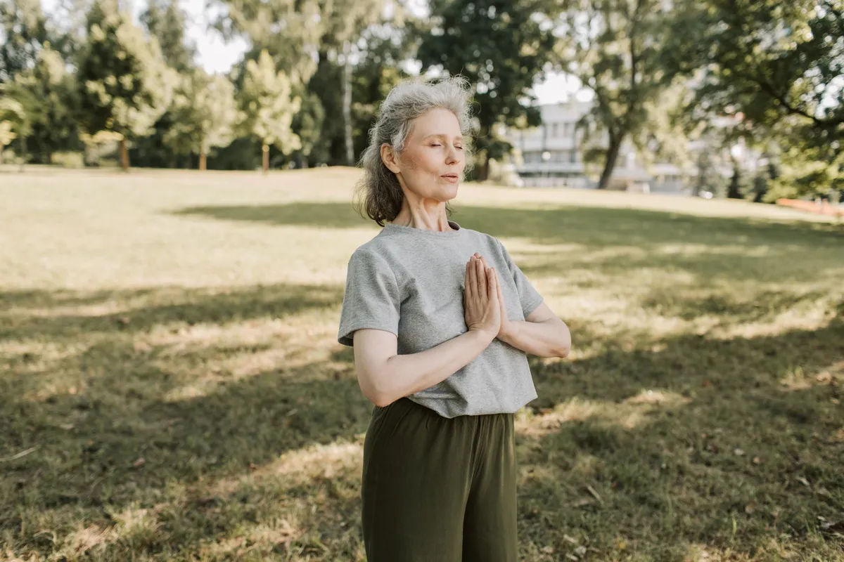 Woman over 50 standing in a sunny park with her hands together, taking a slow breath