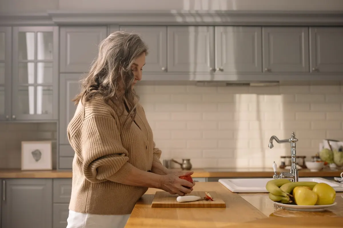 Woman over 50 with gray hair slicing a red pepper on a wooden board in her bright kitchen