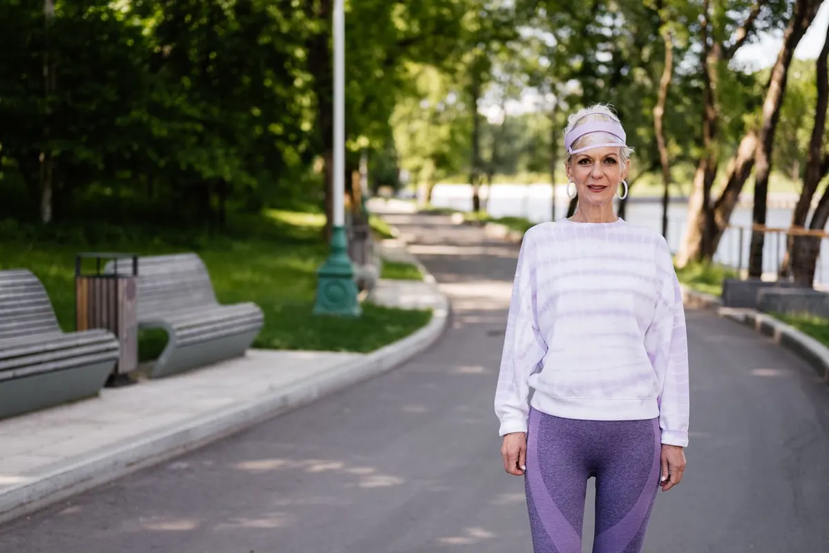 Woman over 50 in a lavender sweatshirt and leggings walking along a sunny tree-lined park path