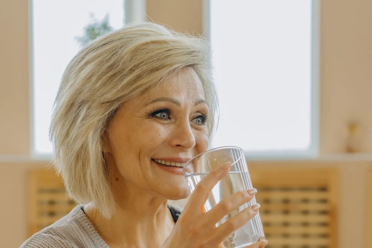 Smiling woman over 50 with a blond bob drinking a glass of water in a bright room