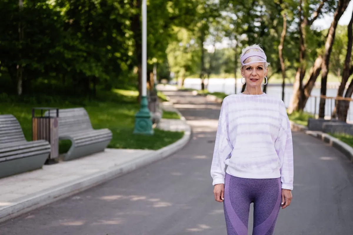 Woman over 50 with gray hair in a lilac sweater and leggings standing on a tree-lined park path