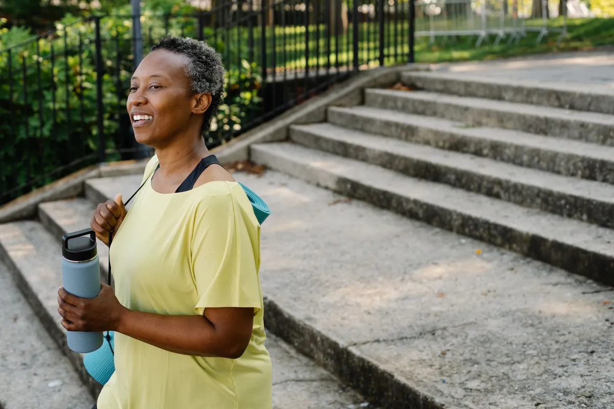Smiling senior woman with short gray hair walking outdoors in a yellow top, carrying a water bottle