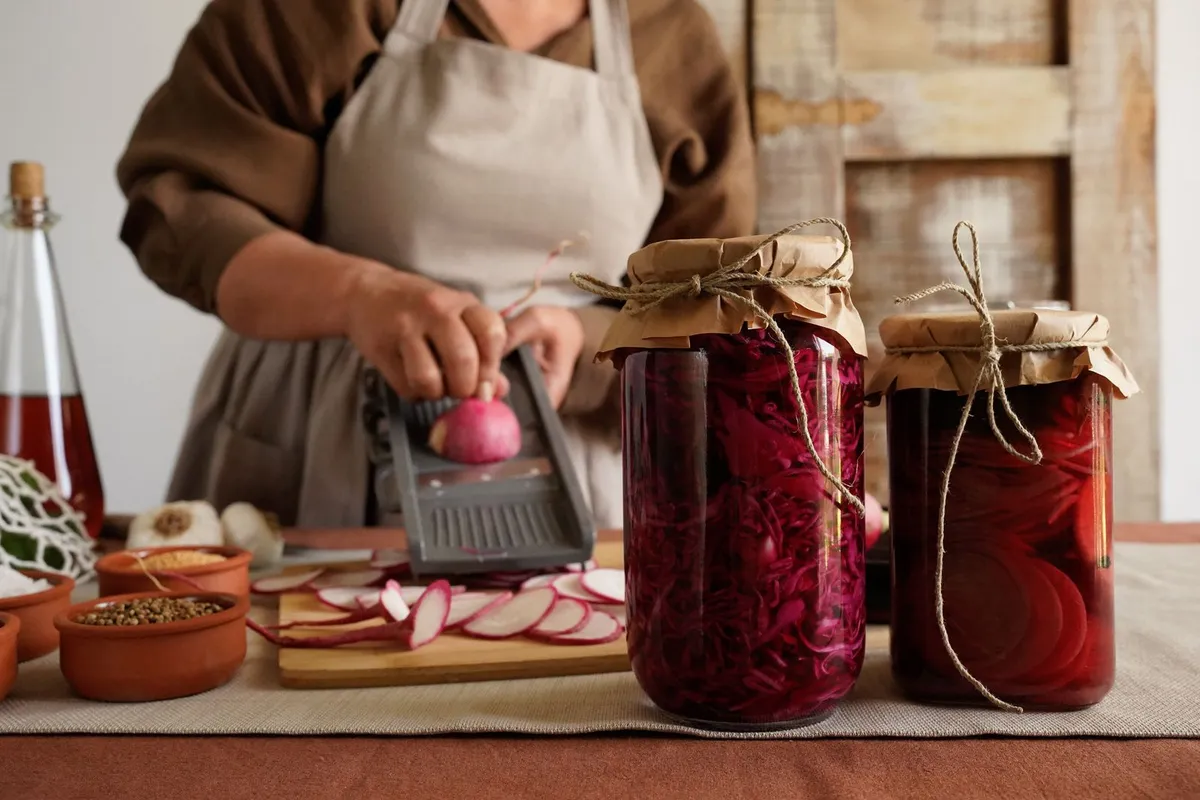 Woman in a linen apron slicing radishes next to two jars of homemade fermented vegetables on a kitchen counter