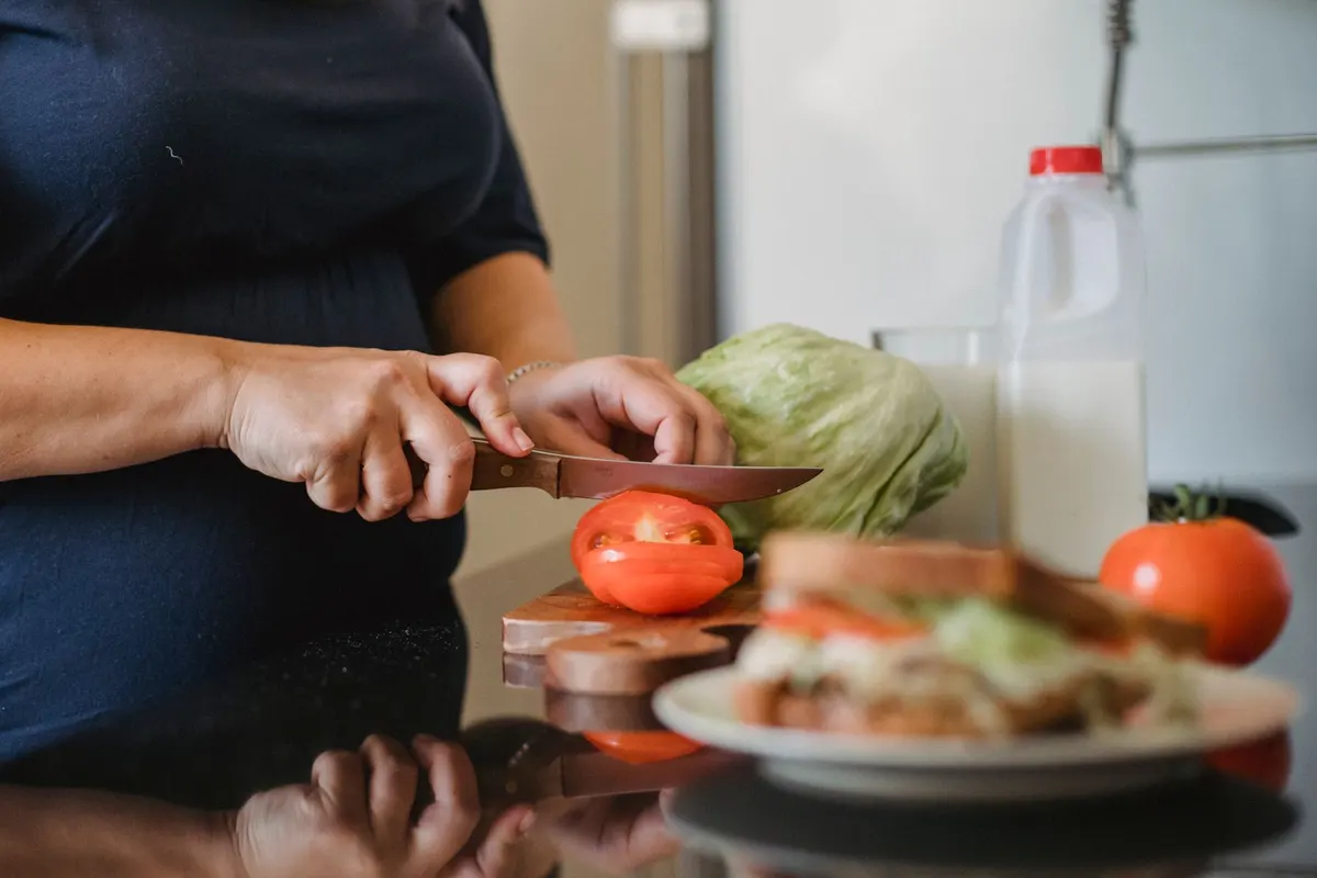 Woman slicing a tomato on a wooden board next to lettuce and a fresh sandwich in a bright kitchen
