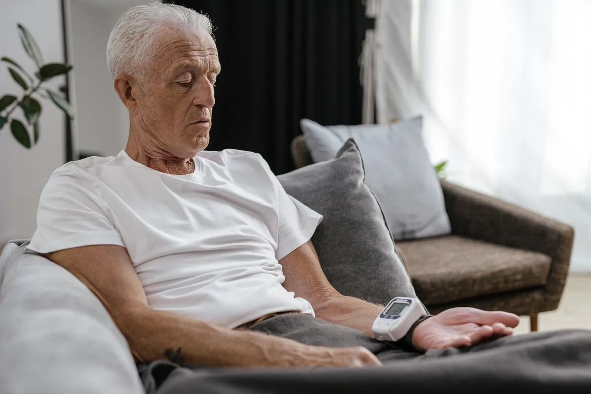 Older man with gray hair resting on a sofa with a wrist blood pressure monitor on his wrist