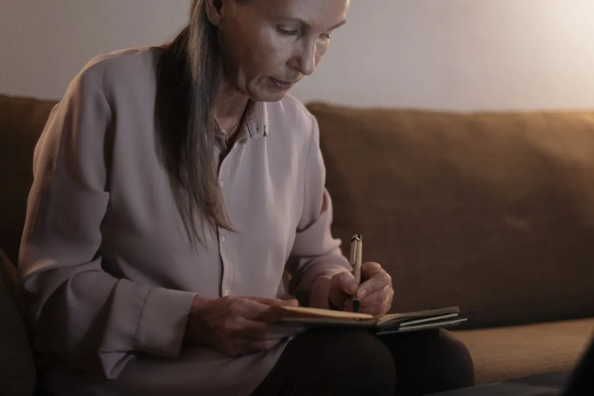 Older woman with long gray hair sitting on a couch and writing in a small notebook