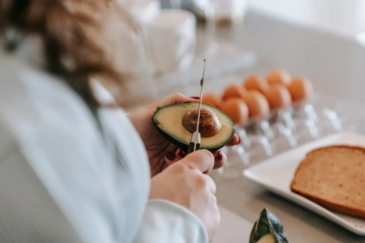 Woman cutting a ripe avocado next to eggs and whole-grain toast in a bright kitchen