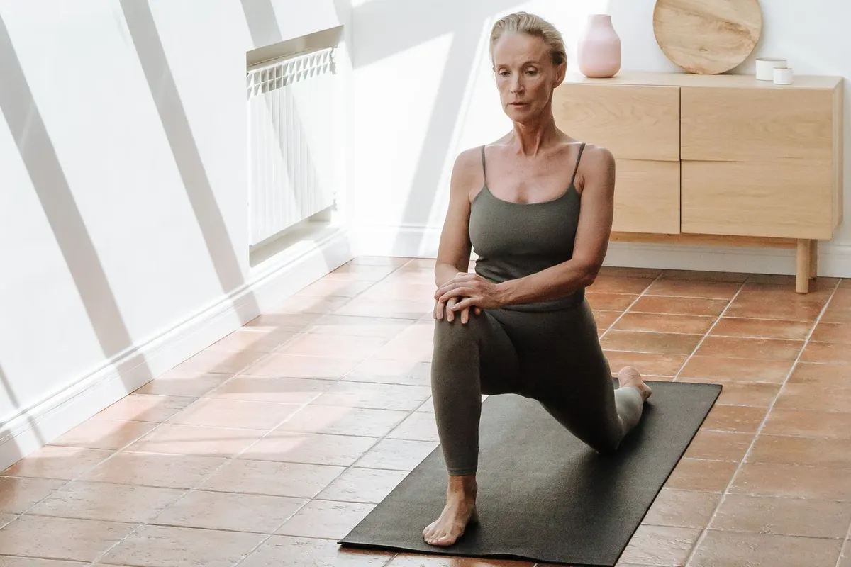 Woman over 50 in a gentle low lunge stretch on a yoga mat in a bright room