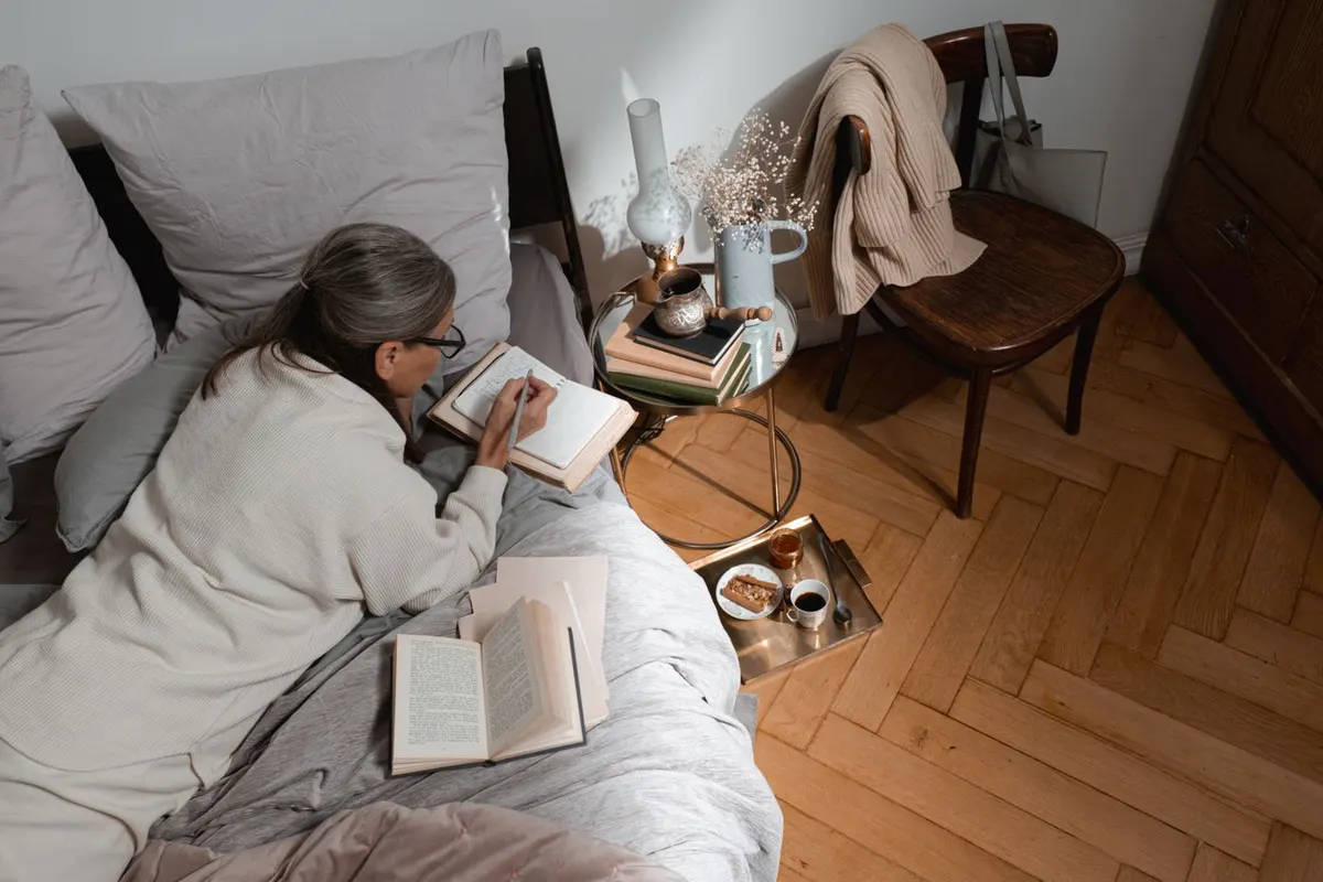 Woman with gray hair resting on a bed with an open book, a cup of tea and a bedside table nearby