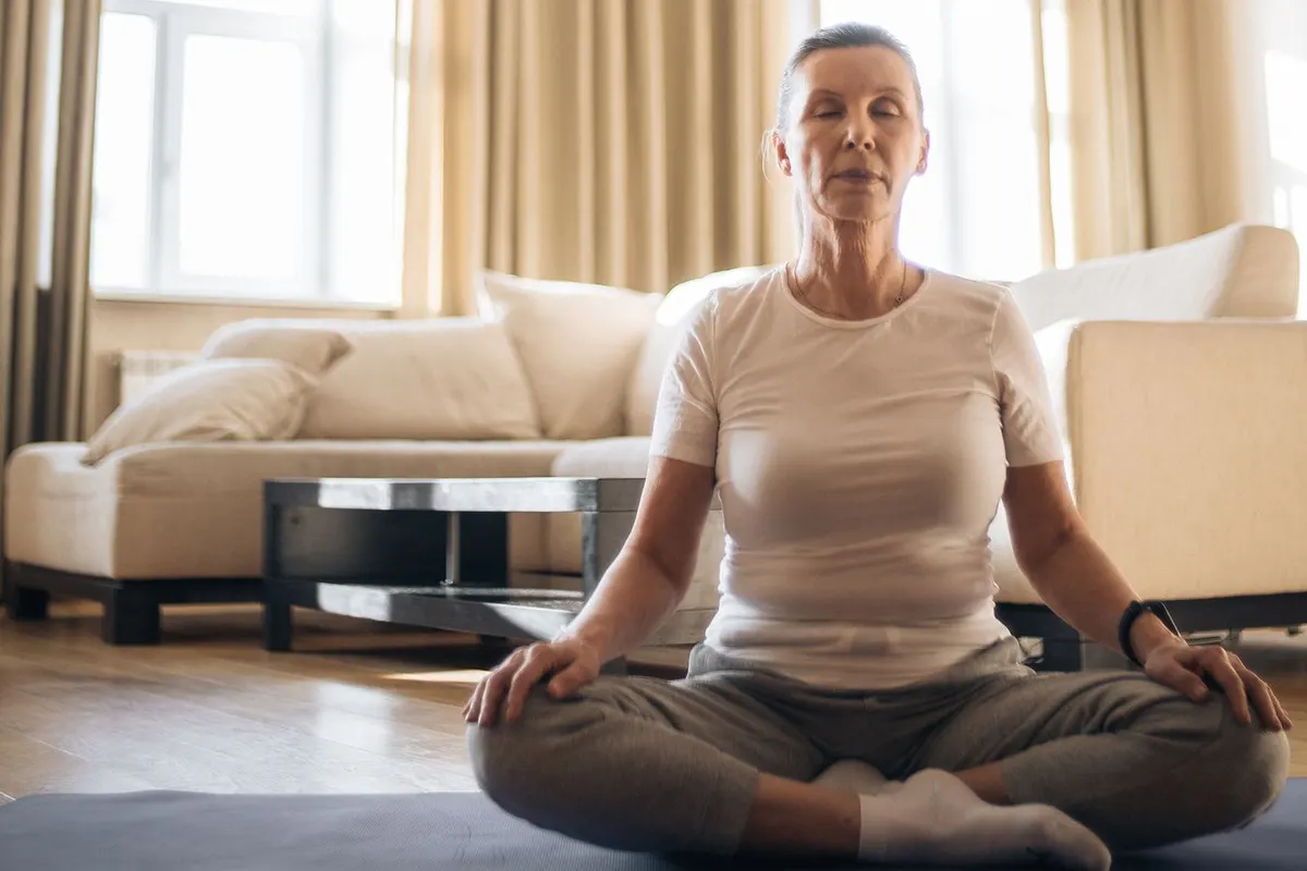 Woman in her 50s sitting cross-legged on a mat in a bright living room with her eyes closed