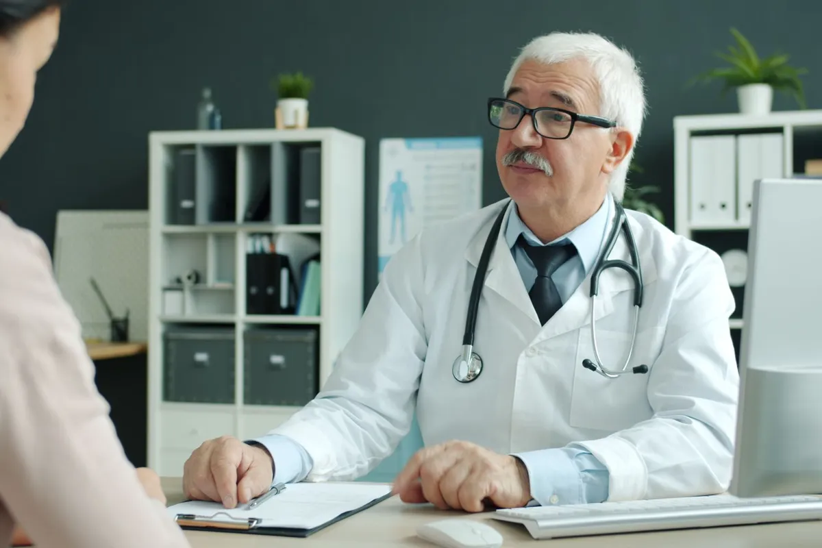 Doctor with gray hair and glasses talking with a patient across a desk in a medical office