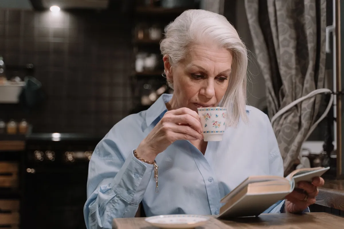 Older woman with gray hair sipping tea from a floral cup while reading a book at a kitchen table
