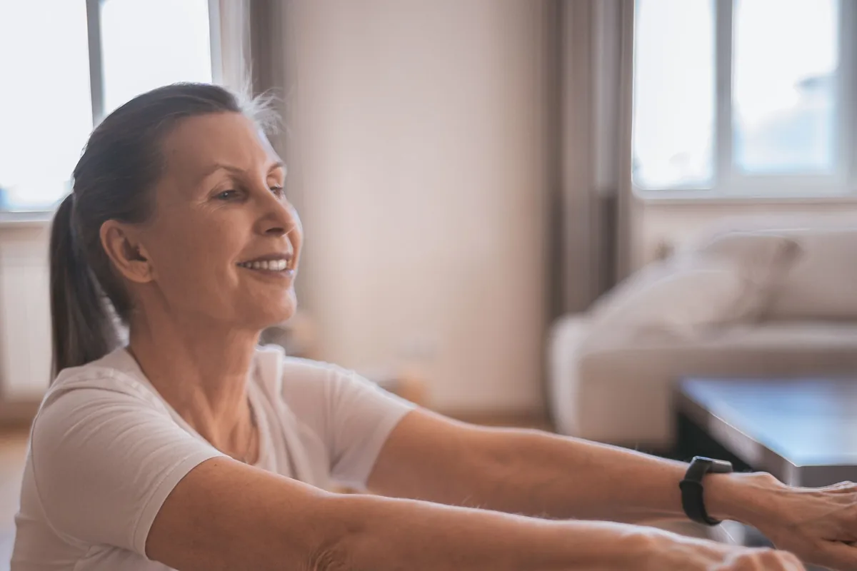 Smiling woman over 50 reaching both arms forward in a seated stretch on a mat at home