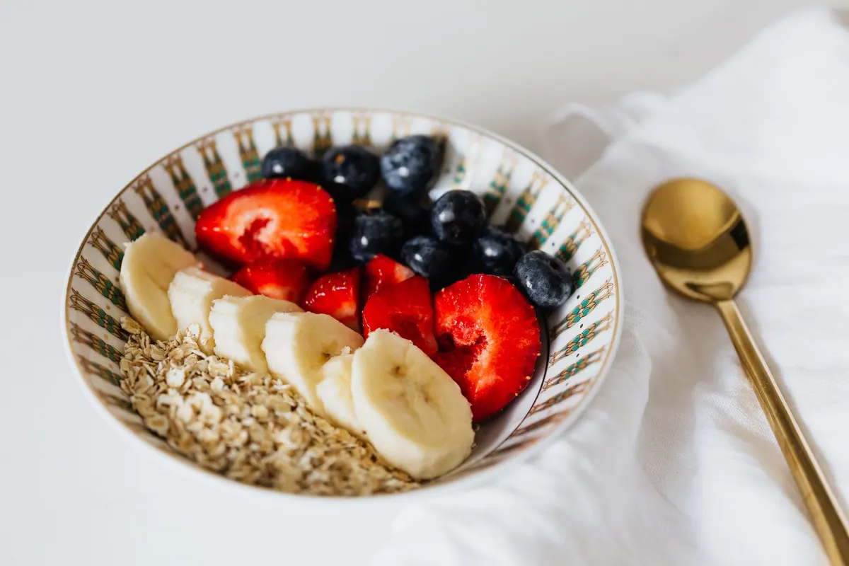 Bowl of oats topped with sliced banana, strawberries and blueberries next to a golden spoon