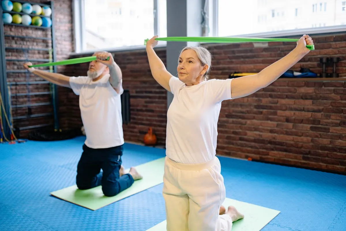Older woman with gray hair stretching a green resistance band above her head on an exercise mat