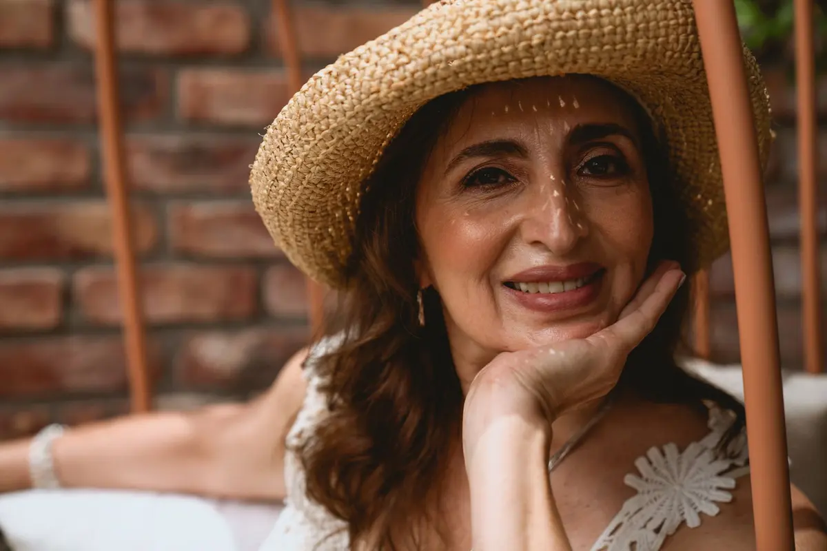 Smiling woman over 50 wearing a straw sun hat in front of a brick wall, resting her chin on her hand