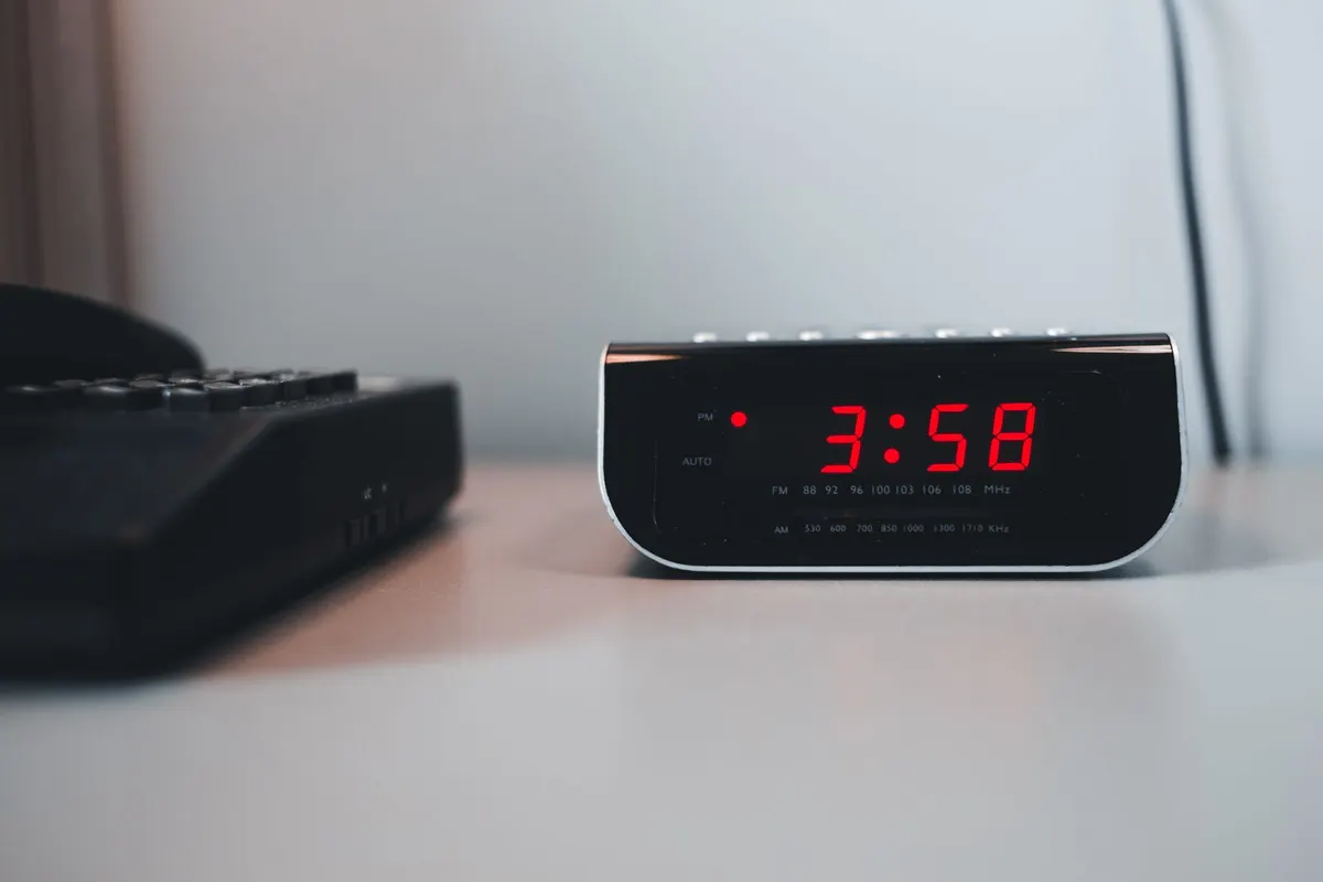Black bedside alarm clock with red digits showing 3:58 on a white nightstand
