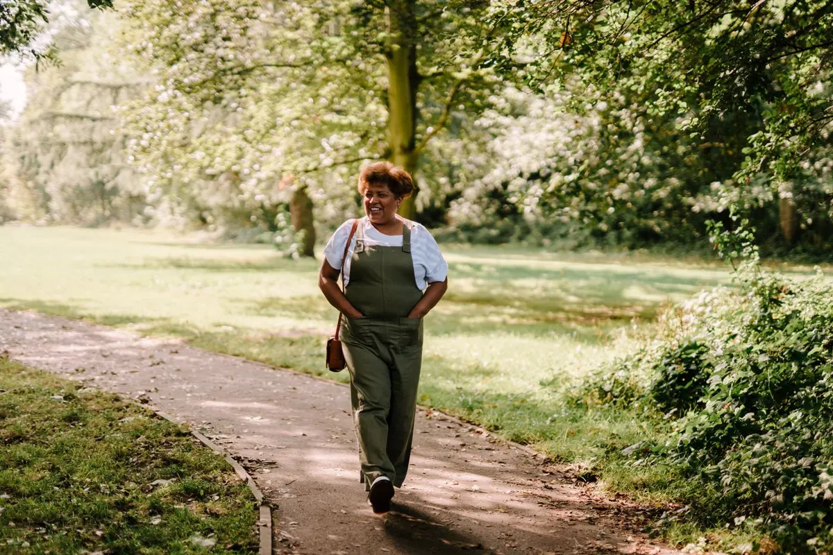 Woman in overalls walking along a tree-lined park path on a sunny day