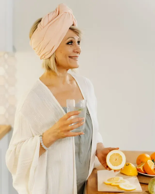 Smiling woman in her fifties holding a glass of water in a bright kitchen next to fresh citrus