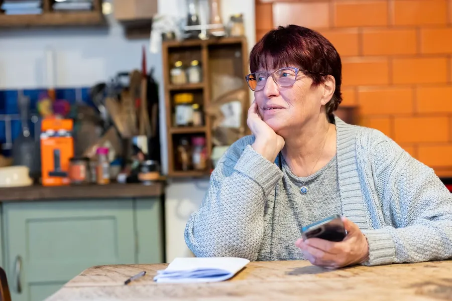 Woman in her sixties sitting at her kitchen table with a notebook, pausing to think