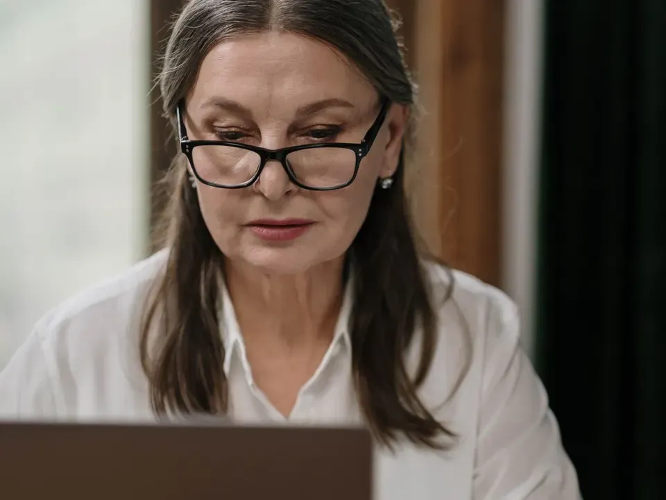 Woman in her fifties with glasses concentrating on her laptop at a wooden table