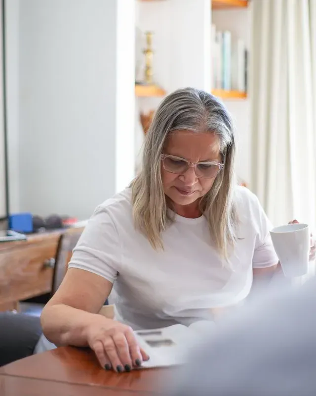 Woman in her fifties with glasses reading at a table and holding a white mug in the morning