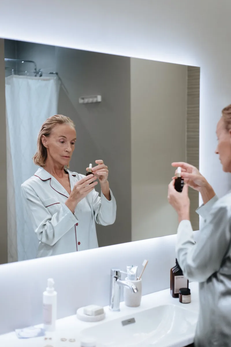 Woman over 50 in pajamas applying a skincare serum while looking in the bathroom mirror