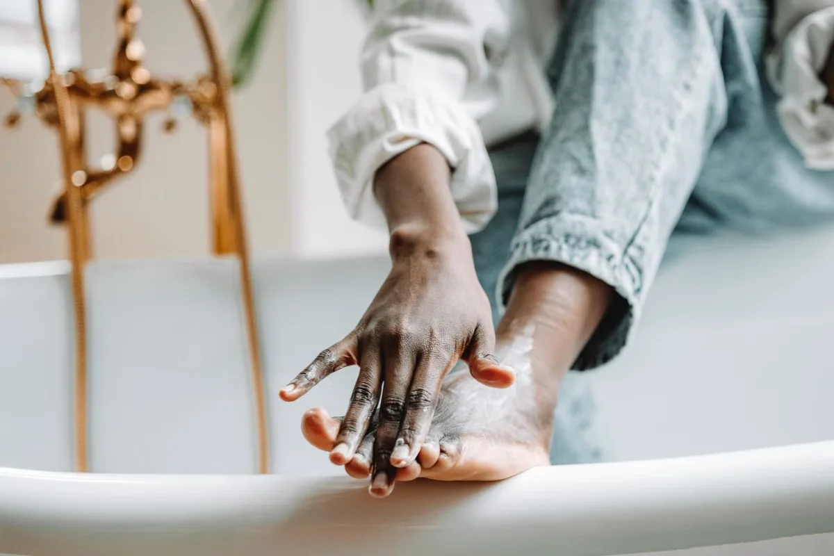 Person sitting on the edge of a bathtub massaging a white cream into the top of a bare foot