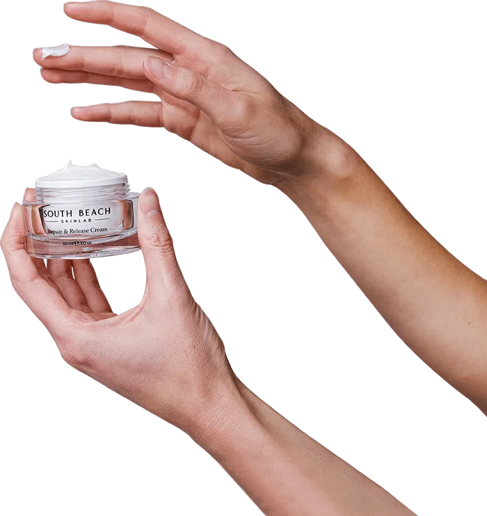 Hand holding an open jar of Repair & Release Cream while the other hand shows a small dab of cream on a fingertip