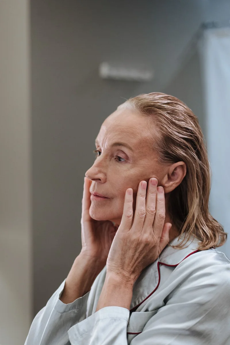 Woman over 50 in pajamas touching her cheeks as she looks at her skin in the bathroom